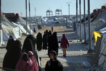 Women and children walk through the Al-Hol Camp in Syria, a displacement site housing over 30,000 people, with tents and infrastructure visible under a clear sky.