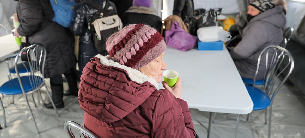 Пожилая женщина в зимней шляпе и пальто выпивает из чашки в временном приюте в Бориспиле (Украина), когда другие люди собираются на фоне в экстремальных зимних условиях.