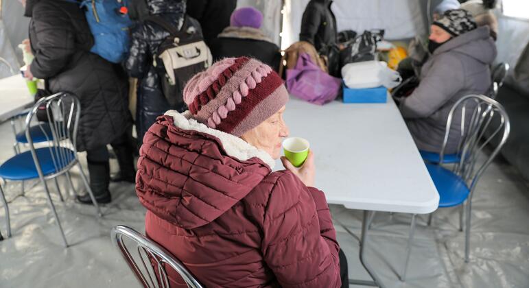 Пожилая женщина в зимней шляпе и пальто выпивает из чашки в временном приюте в Бориспиле (Украина), когда другие люди собираются на фоне в экстремальных зимних условиях.