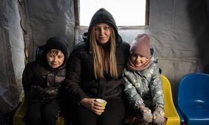 A mother and her two children sit together inside a tent at a heating point in Boryspil, Ukraine, on January 14, 2026. They are warming up and charging devices, as their home lacks electricity for hours. The image highlights the humanitarian crisis affecting displaced families.