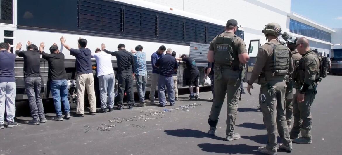 Detained workers with hands raised stand in front of a bus as ICE agents supervise.