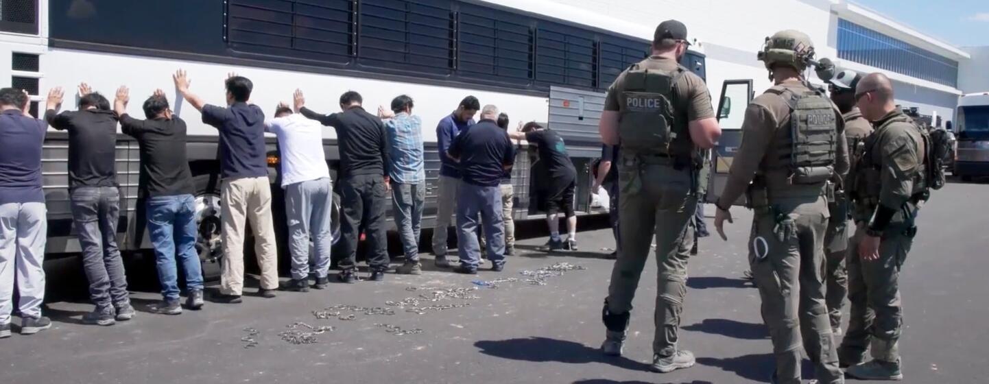 Les travailleurs détenus avec les mains levées se tiennent devant un bus alors que les agents de l'ICE surveillent les installations Hyundai-LG en Géorgie.