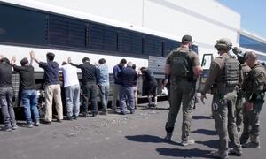 Detained workers with hands raised stand in front of a bus as ICE agents supervise.