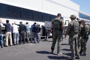 Detained workers with hands raised stand in front of a bus as ICE agents supervise.