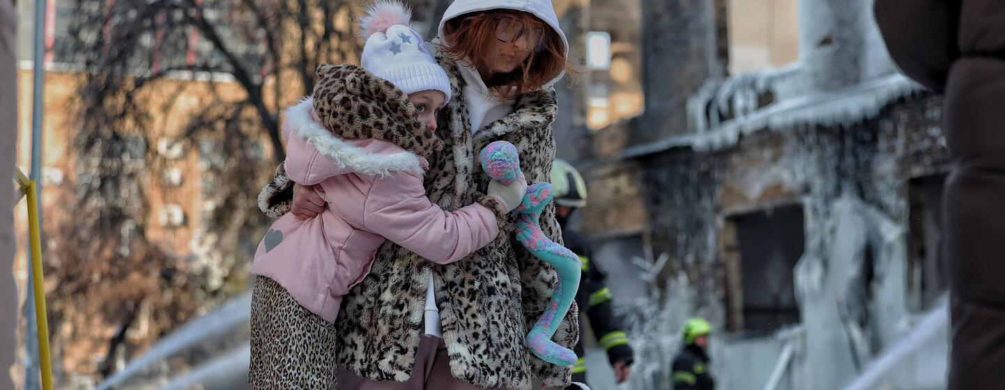 Une mère tient son petit enfant devant un bâtiment lourdement endommagé avec des pompiers pulvérisant de l'eau. La glace recouvre la structure et le sol, soulignant les conditions hivernales difficiles à Kiev, en Ukraine, à la suite d'une attaque contre des infrastructures critiques. L'UNICEF soutient les efforts d'intervention d'urgence pour fournir de la chaleur et des soins essentiels aux familles.