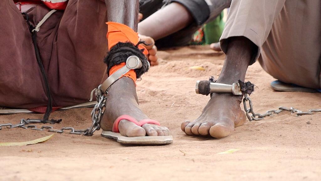 Picha ya karibu ya miguu ya mtu akiwa amefungwa kwa minyororo ya chuma na minyororo, akiwa ameketi kwenye ardhi yenye vumbi nchini Sudan Kusini, ikiashiria kukamatwa na kutafuta haki kupitia mahakama ya mkononi.