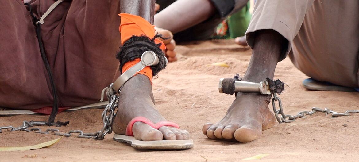 Grand plan des pieds d'une personne enchaînée avec des menottes métalliques et des chaînes, assise sur un sol poussiéreux au Soudan du Sud, symbolisant la détention et la poursuite de la justice à travers un tribunal mobile.