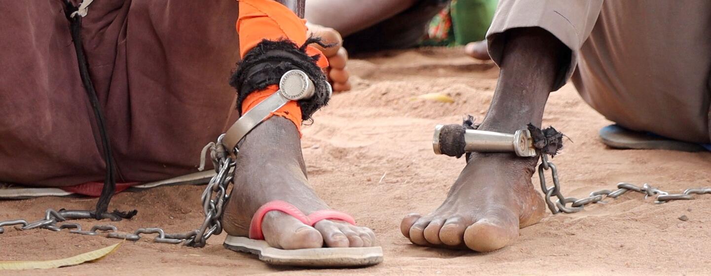 Close-up of a person's feet shackled with metal cuffs and chains, sitting on dusty ground in South Sudan, symbolizing detention and the pursuit of justice through a mobile court.