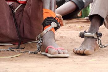 Close-up of a person's feet shackled with metal cuffs and chains, sitting on dusty ground in South Sudan, symbolizing detention and the pursuit of justice through a mobile court.