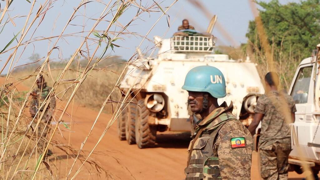 Mwanajeshi wa UNMISS wa kudumisha amani katika kofia ya bluu amesimama kwenye barabara yenye vumbi huko Sudan Kusini, na gari la kivita la kijeshi nyuma. eneo hilo linaonyesha ujumbe wa Umoja wa Mataifa unaounga mkono kupelekwa kwa mahakama ya rununu ili kutoa haki katika maeneo ya mbali.