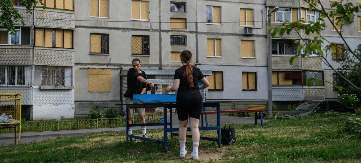 Два человека играют в настольный теннис перед разрушенным войной многоквартирным зданием в Украине с закрытыми окнами.