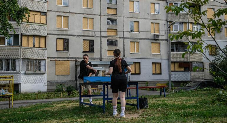 Два человека играют в настольный теннис перед разрушенным войной многоквартирным зданием в Украине с закрытыми окнами.