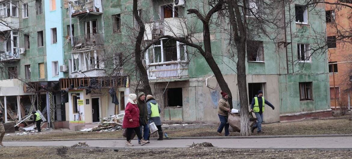 Un edificio de apartamentos de varios pisos dañado en Kramatorsk, Ucrania, con ventanas rotas y escombros. Varios civiles y trabajadores humanitarios con chalecos de alta visibilidad caminan frente al edificio.