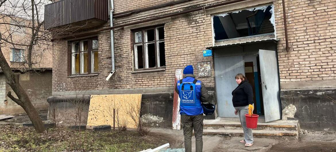 Работник гуманитарной помощи, одетый в синий жилет OCHA, стоит перед поврежденным кирпичным жилым зданием в Крывье-Рихе, Украина, после нападения. Житель стоит рядом, держа красный ведро. Здание демонстрирует значительный ущерб с разбитыми окнами и обломками на земле.