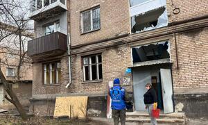 Humanitarian aid worker wearing a blue OCHA vest stands in front of a damaged brick apartment building in Kryvyi Rih, Ukraine, after an attack. A resident stands nearby holding a red bucket. The building shows significant damage with shattered windows and debris on the ground.