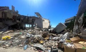 A wide shot of a destroyed building and rubble in Lebanon under a bright blue sky, showing the aftermath of conflict.