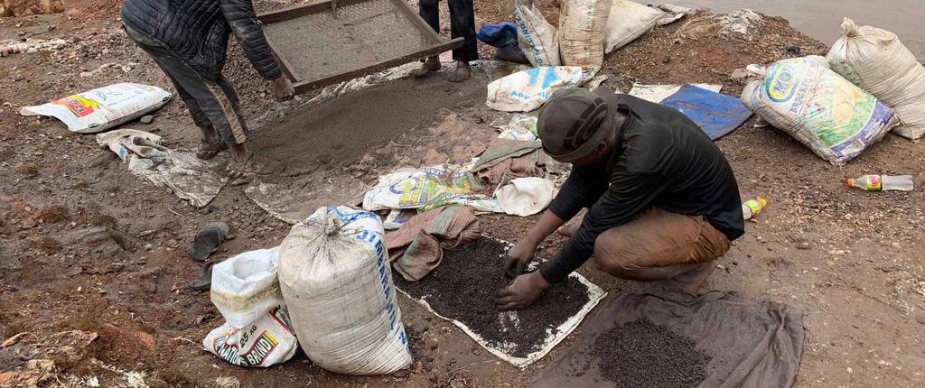 Mwanamume anapiga magoti kwenye ardhi katika mgodi wa madini wa DR Congo, akichunguza udongo mweusi kwa ajili ya cobalt. Mifuko mikubwa ya rangi nyeupe imepangwa kuzunguka.