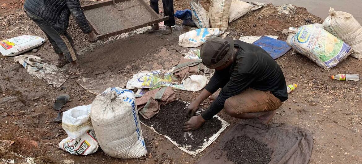 A man crouches on the ground in a pit mine in DR Congo, sifting dark soil for cobalt. Large white sacks are scattered around him.