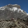 A person on horseback traverses a rugged, rocky trail in the foreground, with a massive, snow-capped mountain dominating the background under a clear blue sky.