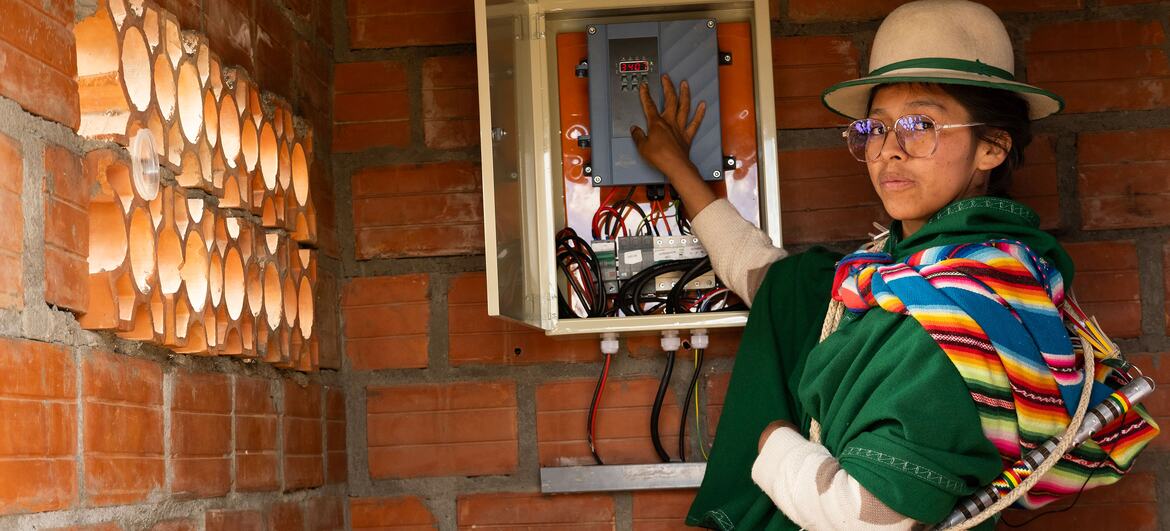 A Bolivian woman in traditional colorful clothing and a hat stands next to an open solar energy control panel mounted on a brick wall, demonstrating the impact of technology on rural communities.