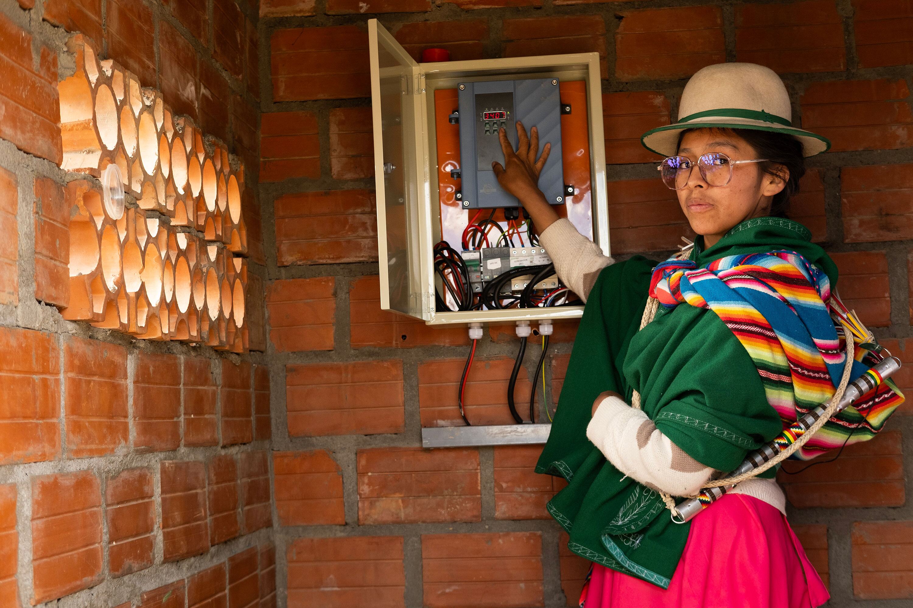 امرأة بوليفية ترتدي ملابس تقليدية ملونة وقبعة تقف بجانب لوحة تحكم للطاقة الشمسية مفتوحة مثبتة على جدار من الطوب، مما يوضح تأثير التكنولوجيا على المجتمعات الريفية.