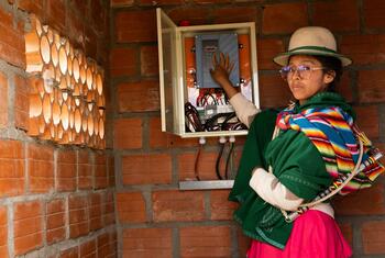 A Bolivian woman in traditional colorful clothing and a hat stands next to an open solar energy control panel mounted on a brick wall, demonstrating the technology's impact on rural communities.
