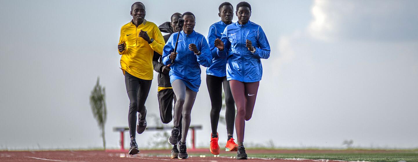 A group of young African athletes, both male and female, running on a red track during a training session at the Teryet National High Altitude Training Centre in Kapchorwa District, Uganda.