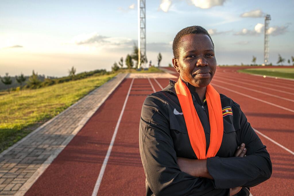 Portrait of a female coach at the Teryet National High Altitude Training Centre in Kapchorwa District, Uganda. She stands on an athletics track with her arms crossed, wearing a black athletic jacket with a Ugandan flag patch and an orange scarf.