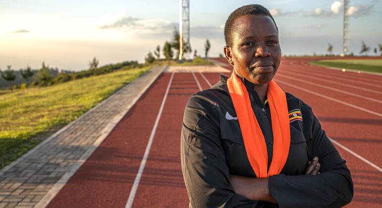 Portrait of a female coach at the Teryet National High Altitude Training Centre in Kapchorwa District, Uganda. She stands on an athletics track with her arms crossed, wearing a black athletic jacket with a Ugandan flag patch and an orange scarf.