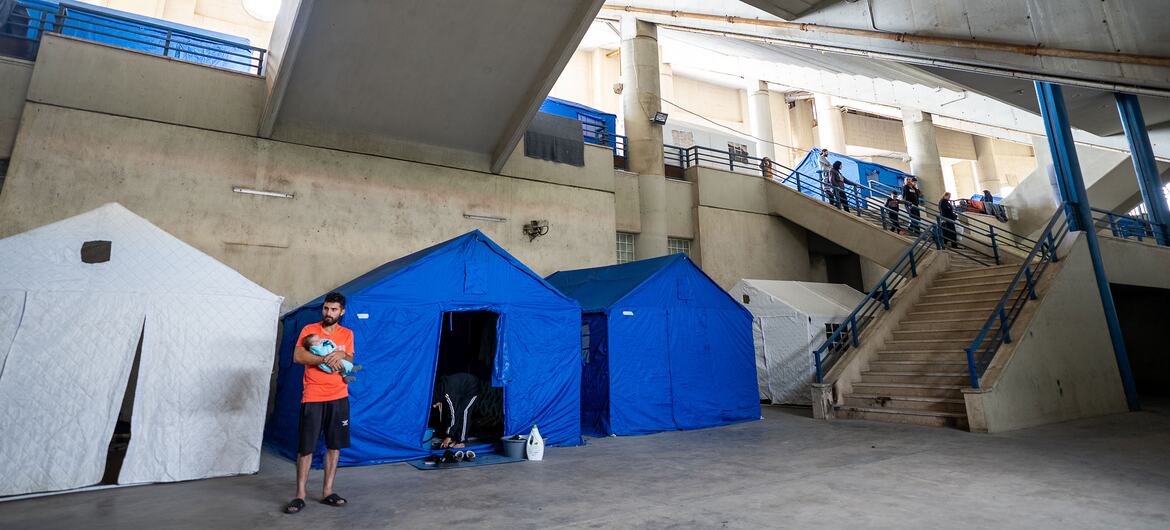Un homme tenant un bébé se tient à l'extérieur d'une tente bleue installée à l'intérieur d'un bâtiment en béton, qui est utilisé comme abri pour les familles déplacées à Beyrouth, au Liban.