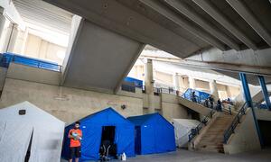 A man holding a baby stands outside a blue tent set up inside a concrete building, which is being used as a shelter for displaced families in Beirut, Lebanon.
