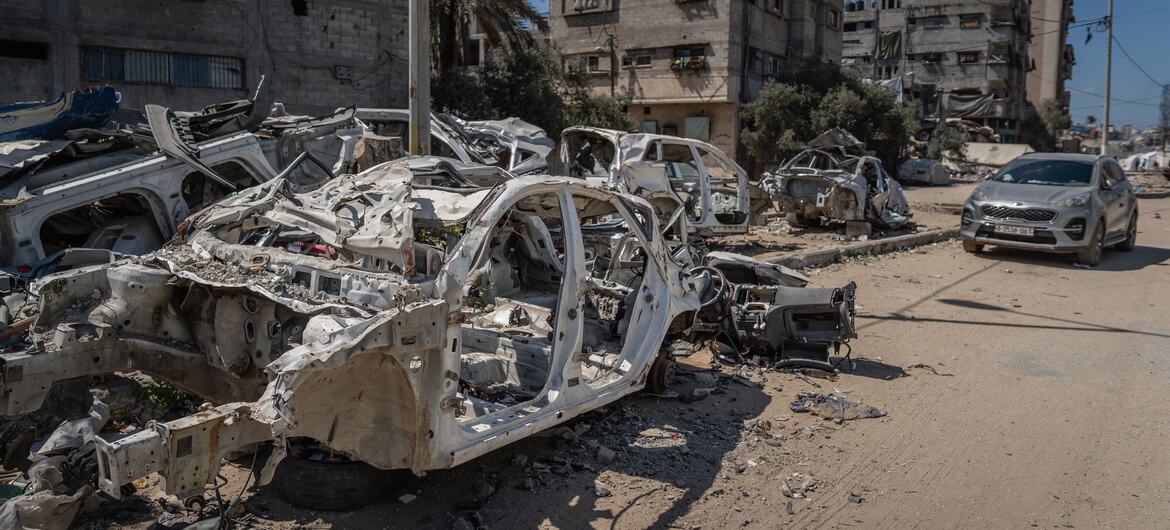 Coches destruidos y quemados en una calle de Gaza, con edificios dañados en el fondo bajo un cielo azul claro, que muestra las consecuencias del conflicto.