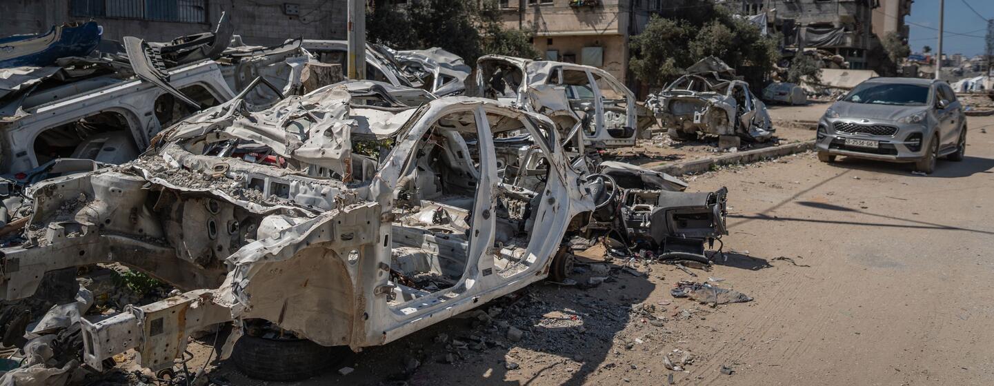 Des voitures détruites et brûlées dans une rue de Gaza, avec des bâtiments endommagés à l'arrière-plan sous un ciel bleu clair, représentant les conséquences du conflit.