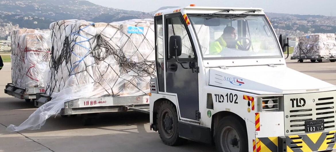 Un vehículo de carga blanco, etiquetado TLD 102, transporta grandes paletas envueltas en plástico y redes a través de una pista del aeropuerto. En el fondo, se ven una ciudad y colinas. Este envío de suministros médicos esenciales fue recibido en el Aeropuerto Internacional Rafik Hariri en el Líbano para apoyar el sistema de salud del país.