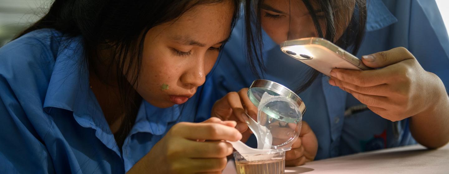 Deux étudiantes en uniforme bleu à l'école Angsilapittayakom de Chonburi, en Thaïlande, inspectent des larves de moustiques à l'aide d'une loupe et d'un smartphone dans le cadre d'une activité d'apprentissage STEM de l'UNICEF.