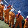 Un groupe de cinq marins en combinaisons orange et casques blancs se tiennent sur le pont d'un navire, en train d'installer du fil de fer barbelé le long de la rambarde, sur fond de mer bleue.