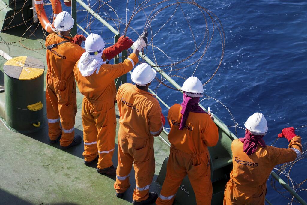 Un groupe de cinq marins en combinaisons orange et casques blancs se tiennent sur le pont d'un navire, en train d'installer du fil de fer barbelé le long de la rambarde, sur fond de mer bleue.