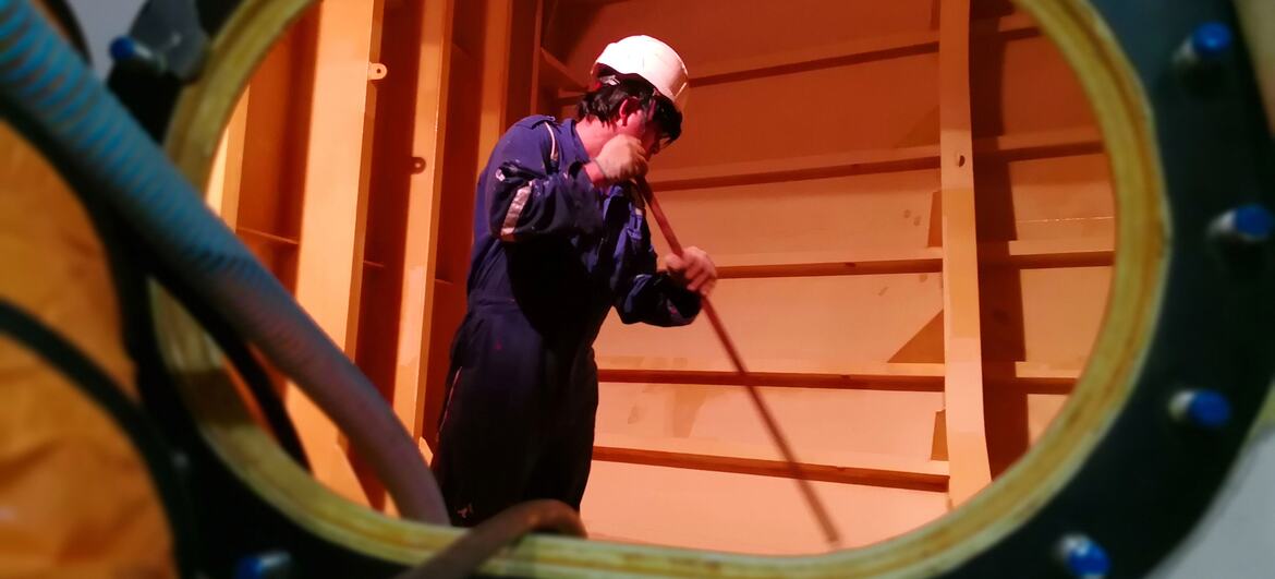 A Filipino seafarer in a blue uniform and white hard hat is seen through a circular porthole, cleaning the interior of a ship's cargo hold or warehouse.
