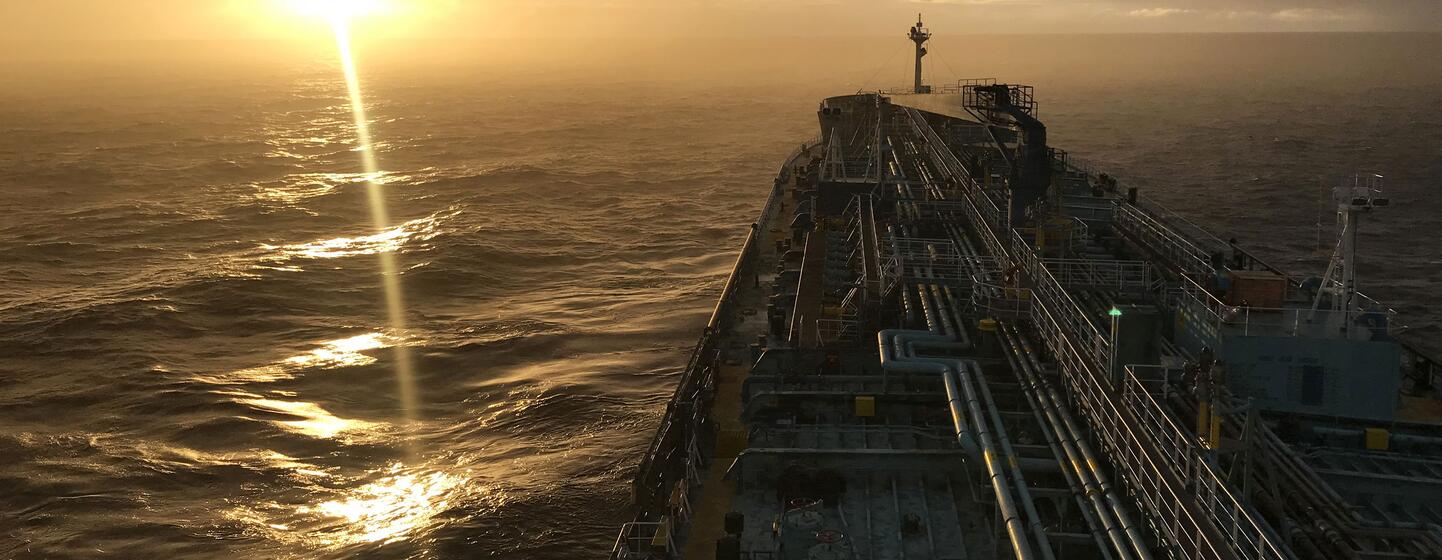 Vista do convés de um grande navio de carga ou petroleiro navegando no mar aberto durante o pôr do sol. O sol está se pondo no horizonte, lançando um brilho dourado sobre a água e o convés do navio.