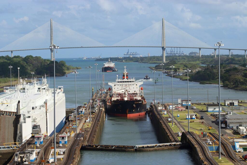 A container ship navigates through the Panama Canal, passing under a large white bridge with industrial port structures visible in the background.