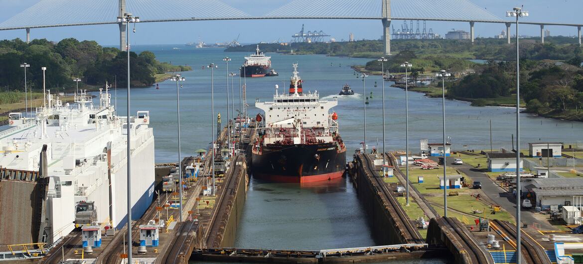 Контейнеровоз, идущий через Панамский канал и проходящий под большим белым мостом, на заднем плане видны промышленные портовые сооружения.