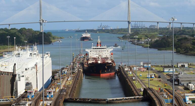 A container ship navigating through the Panama Canal, passing under a large white bridge with industrial port structures visible in the background.