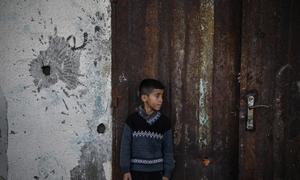A young boy stands by a wall showing the signs of a missile attack.