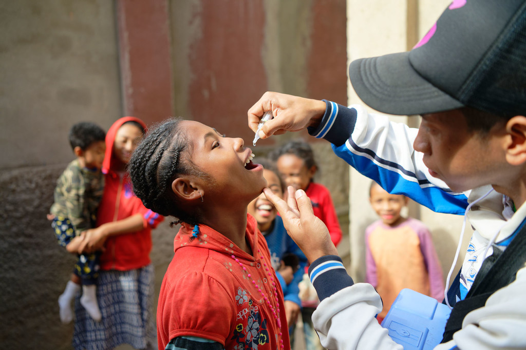 Mtoa chanjo wa kiume akimpa msichana mdogo chanjo ya polio nchini Madagaska kama sehemu ya kampeni ya afya ya jamii. Watoto wengine na wanajamii wanatazama nyuma.