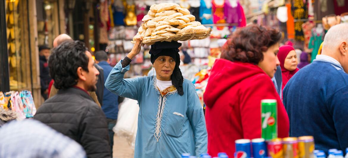 A woman in a blue dress balances a large basket of bread on her head while walking through a bustling market in Cairo, Egypt.