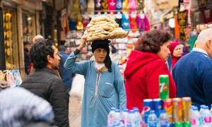 A woman in a blue dress balances a large basket of bread on her head while walking through a bustling market in Cairo, Egypt.
