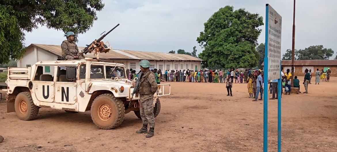 Un véhicule de maintien de la paix de l'ONU avec du personnel armé est stationné dans une zone poussiéreuse de la République centrafricaine, avec une foule de personnes locales rassemblées à l'arrière-plan.