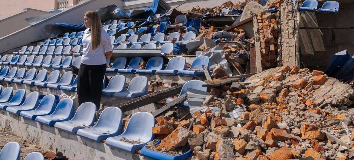 A 17-year-old Ukrainian student named Sofiia stands among the destroyed seats of a stadium in Kharkiv, Ukraine. The grandstand was damaged by shelling, with rubble and broken plastic chairs scattered around. Sofiia, who dreams of becoming a doctor, is part of a UNICEF-supported learning recovery program helping students catch up on education disrupted by the war.