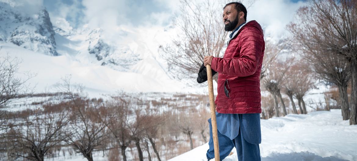 Um homem com jaqueta vermelha e shalwar kameez azul está de pé na neve segurando uma pá, com as montanhas cobertas de neve do Vale de Chunda ao fundo.