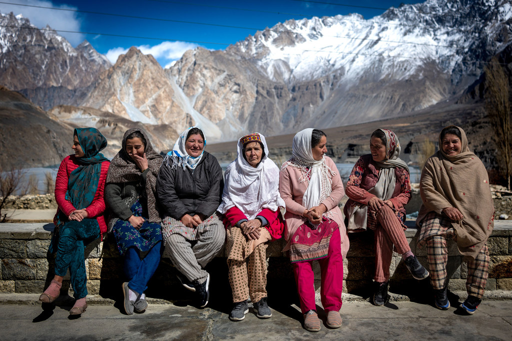 पारंपरिक पोशाक पहने महिलाओं का एक समूह हुंजा घाटी के पहाड़ों और ग्लेशियरों की पृष्ठभूमि में एक पत्थर की दीवार पर बैठा है।